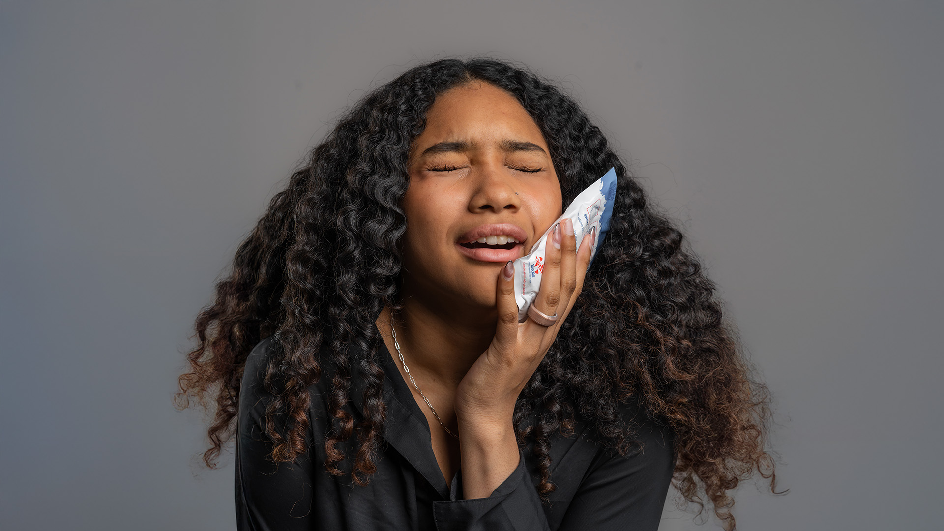 A woman with curly hair is holding her hand over her mouth, appearing surprised or shocked, with a blue object near her face.