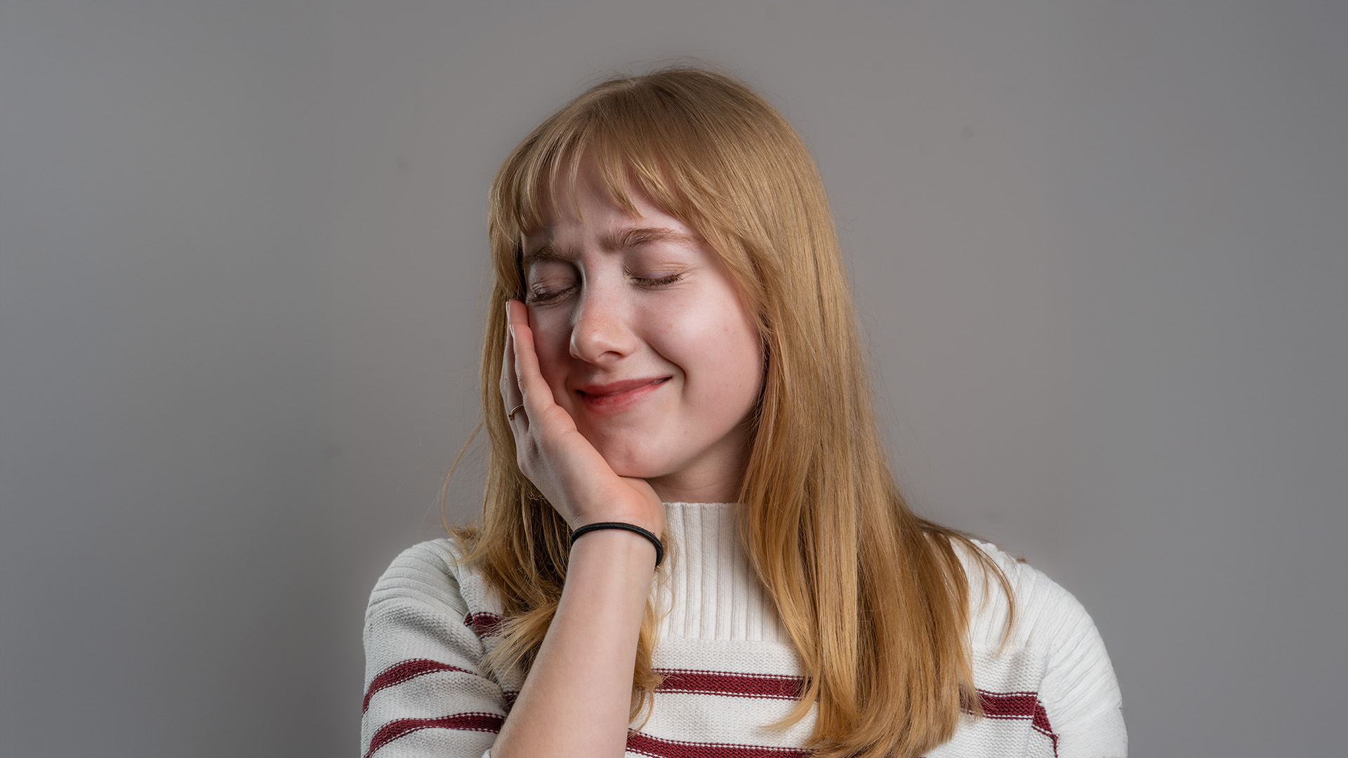 A young woman with blonde hair, wearing a white sweater with horizontal stripes, has her hand on her face with a thoughtful expression.