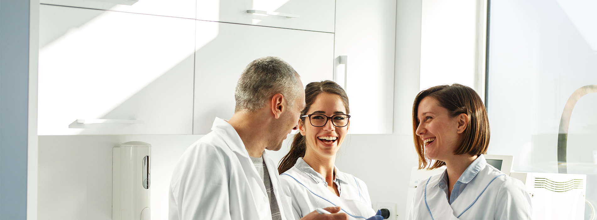 Three individuals are standing in a professional setting, possibly a medical or dental office, with one person holding a tablet, all smiling and engaged in conversation.